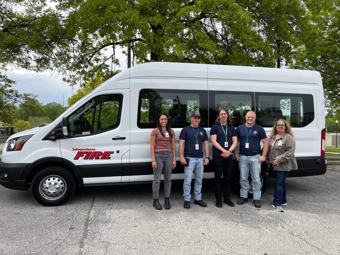 Members of the Independence ARCH program stand next to their large 15 passenger white van parked in front of green trees. 
