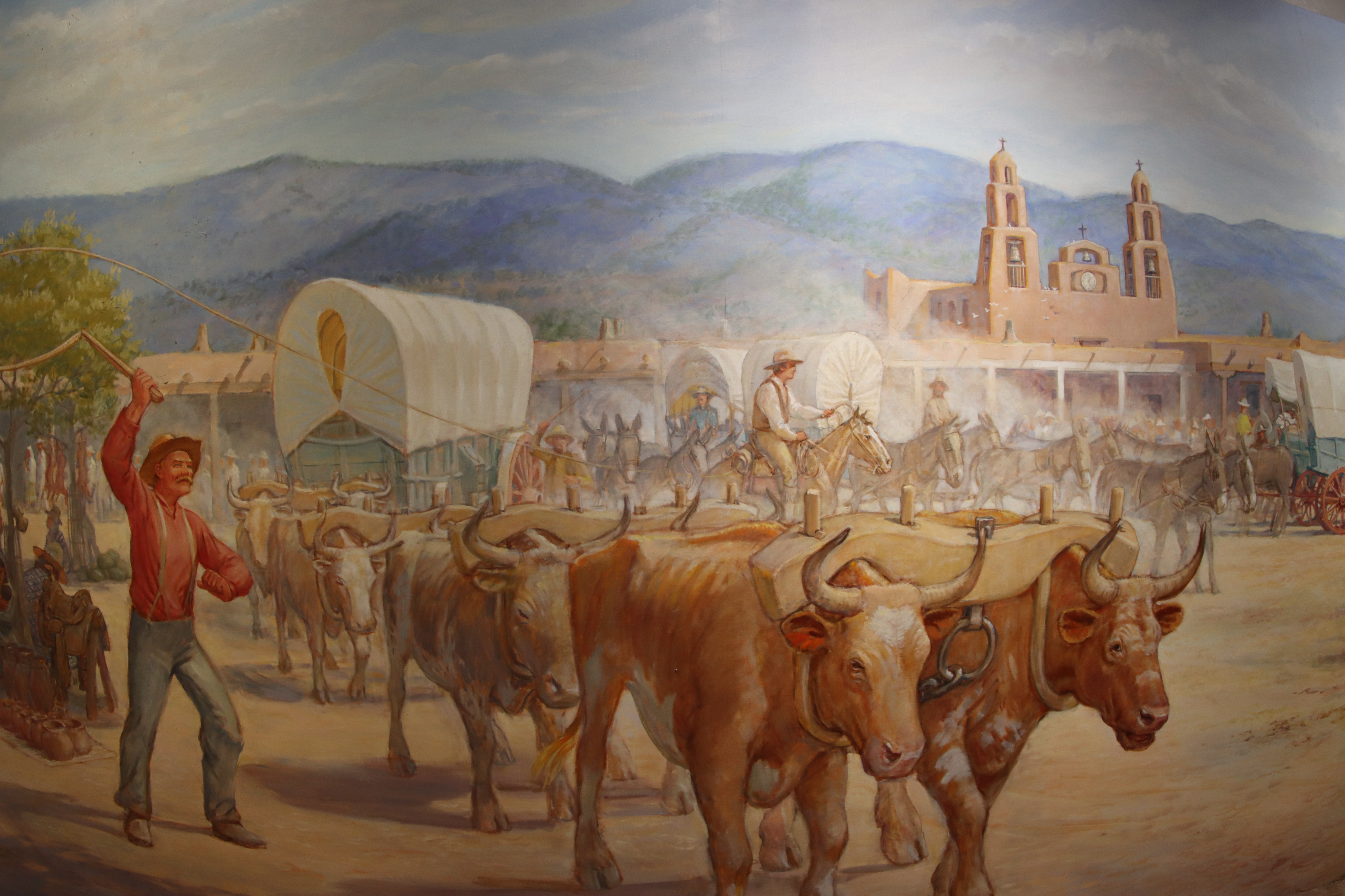 Image of a mural depicting pioneers heading west with oxen pulling a covered wagon on a dirt road and activity behind them. 