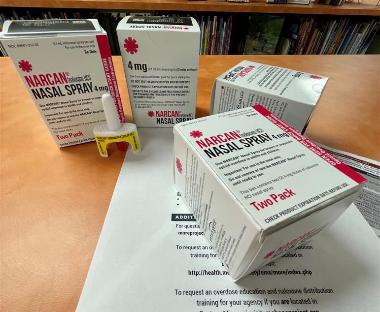 A box of Narcan, a small white bottle with a yellow base, sits on top of a pile of papers discussing drug addiction and treatment options