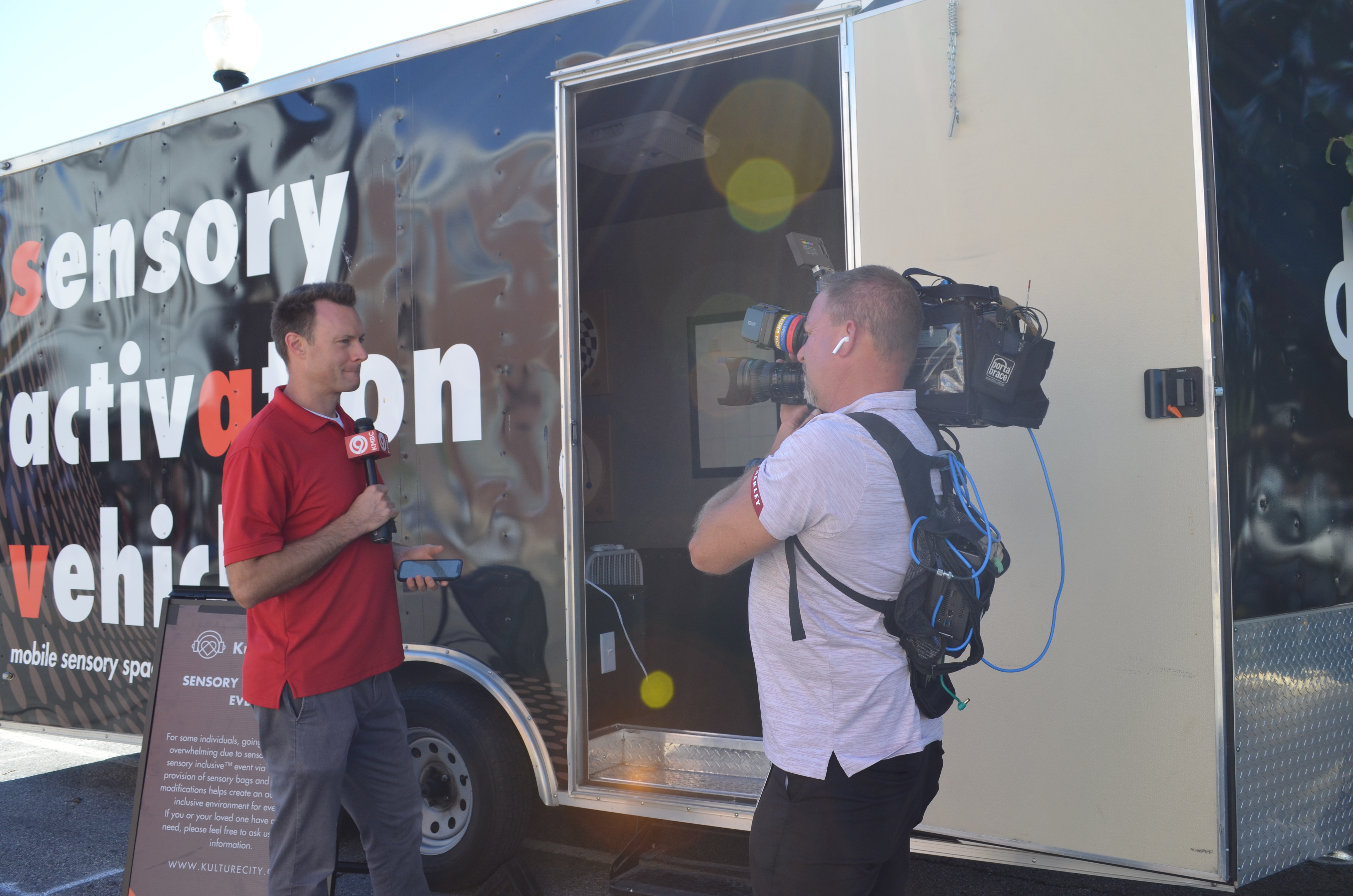 A reporter in a red shirt stands outside of the sensory activation trailer, a black van