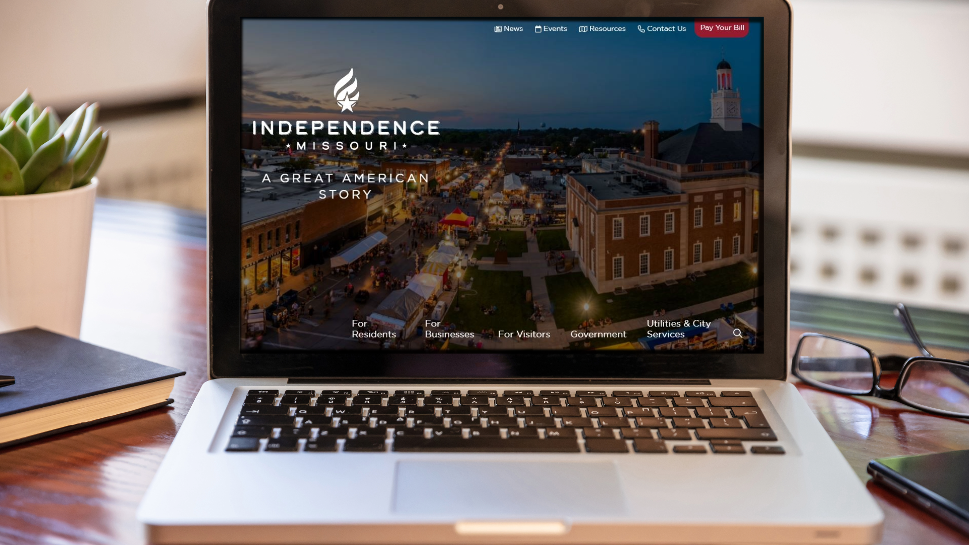 A silver laptop sits on a desk with a plant, a book, a phone, and glasses beside it. The screen shows the new City of Independence website. 