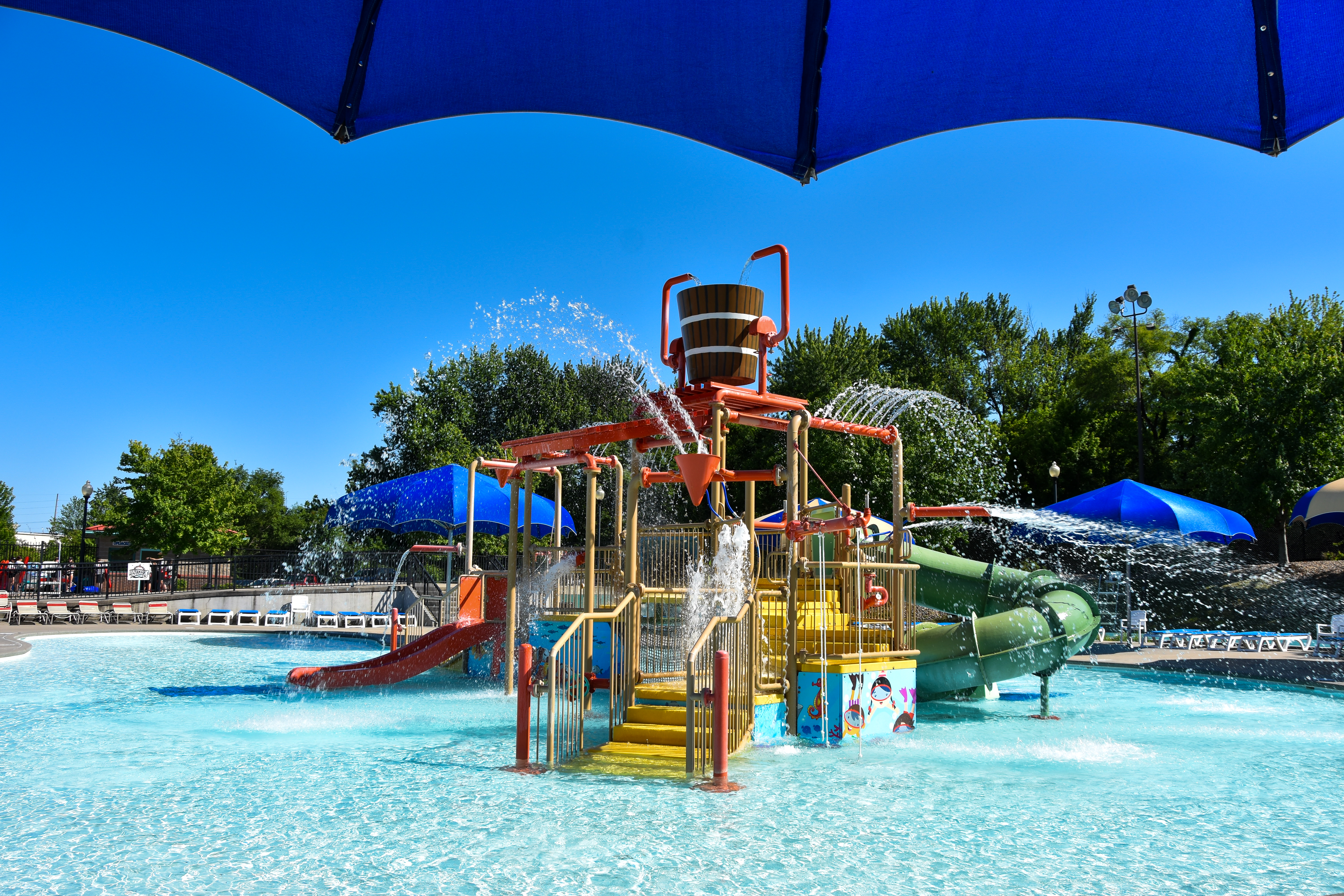 A water park filled with sparkly blue water and a clear blue sky. The water park equipment is brightly painted in a rainbow of colors