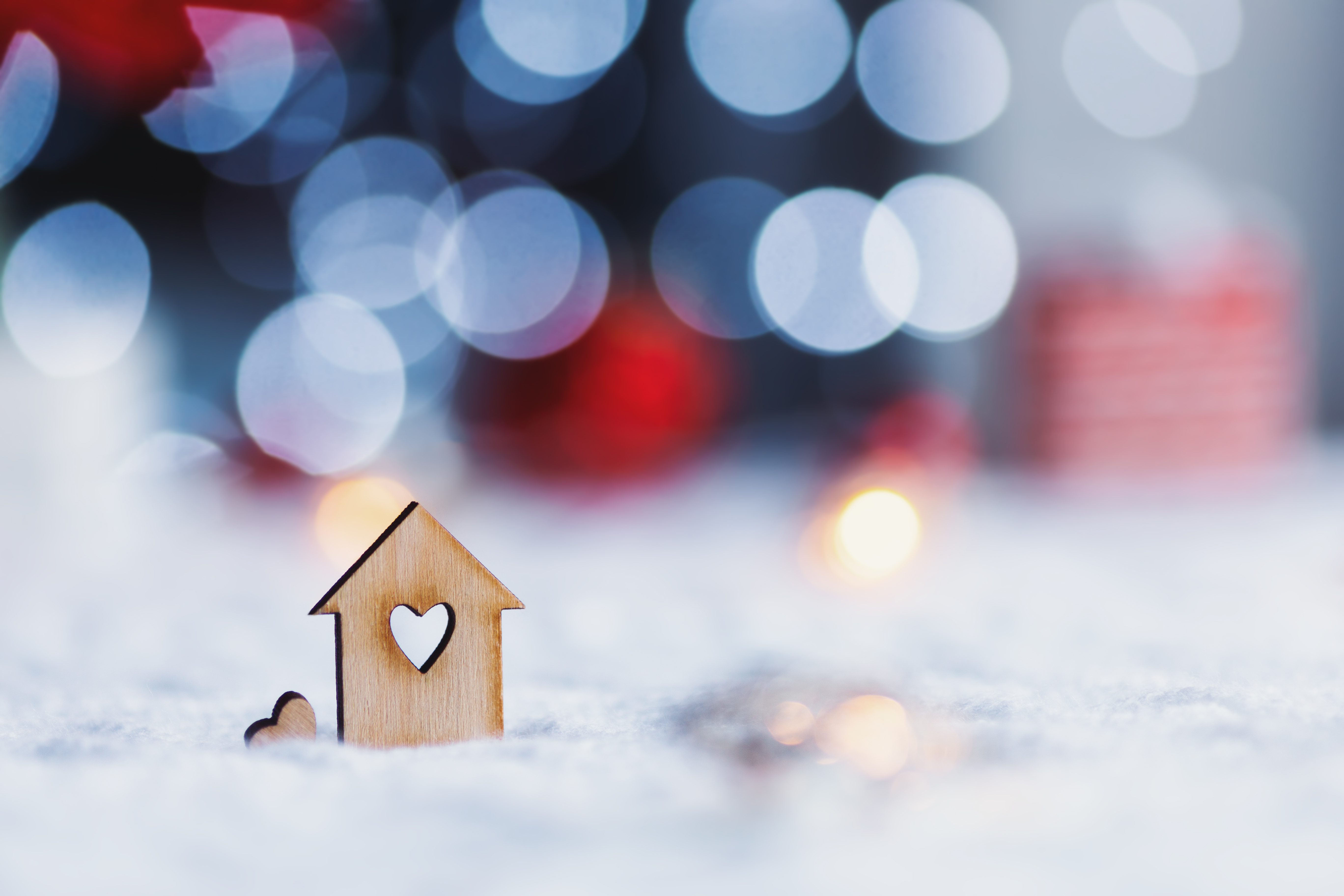 A small wooden home cutout sits in the snow with a heart cut out of the middle of it. Holiday lights and gifts are blurred in the background.