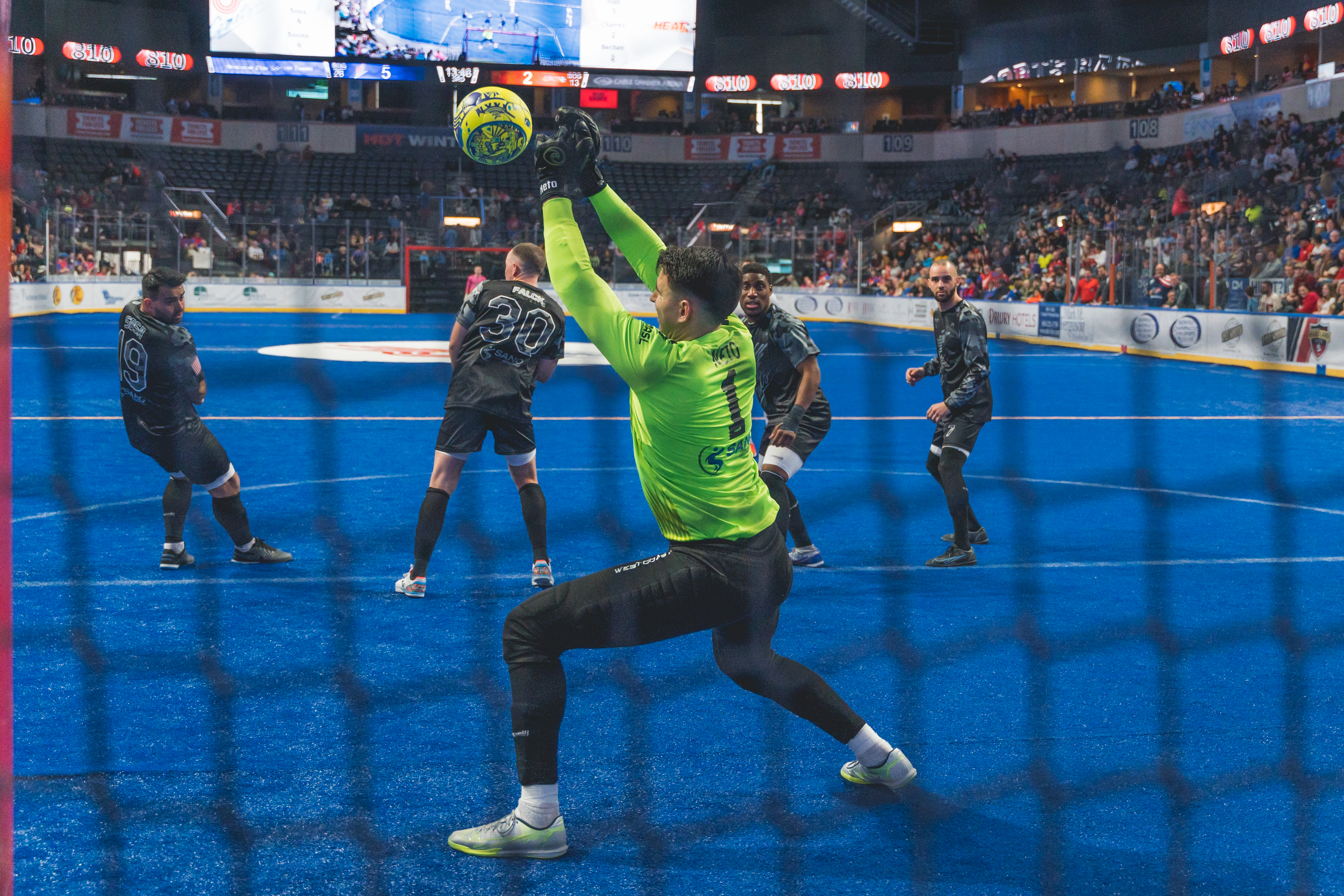 A soccer goalie in a bright yellow vest blocks a soccer ball heading for the goal