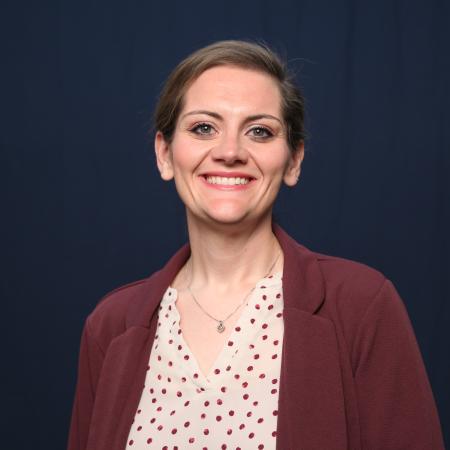 A portrait image of Samantha Morris in a maroon jacket with a peach and maroon shirt under it in front of a dark blue background.