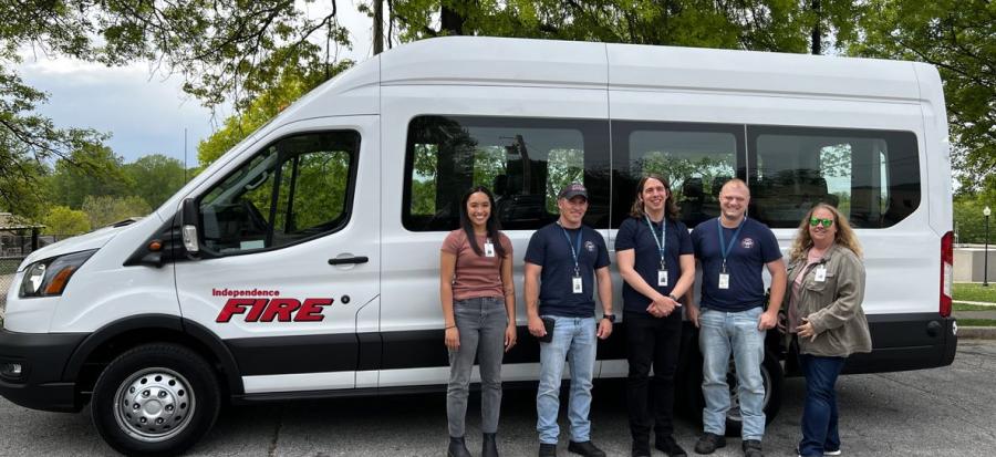 Members of the Independence ARCH program stand next to their large 15 passenger white van parked in front of green trees. 