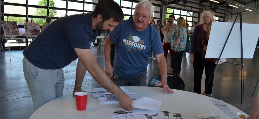 A project team member explains details of the Truman Connected and Downtown Streetscape projects to a member of the public in front of a round table at the Independence Uptown Market. 