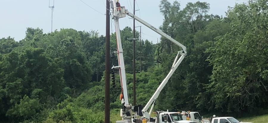 A journymen linemen repairs a powerline from the bucket of an Independence Power and Light bucket truck with damaged trees and grey skies behind them. 