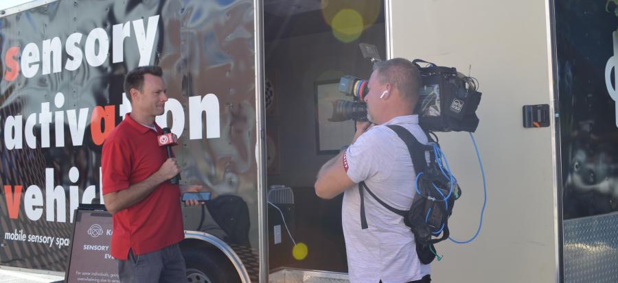 A reporter in a red shirt stands outside of the sensory activation trailer, a black van