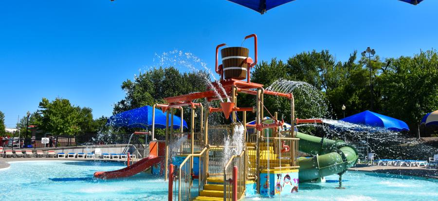 A water park filled with sparkly blue water and a clear blue sky. The water park equipment is brightly painted in a rainbow of colors