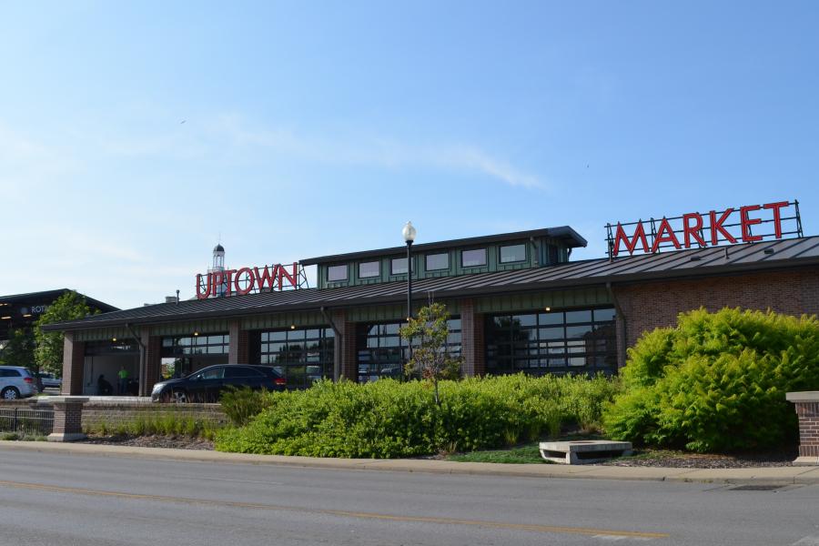 An image of the north side of the Independence Uptown Market building during the daytime with a few parked vehicles and some green landscape.