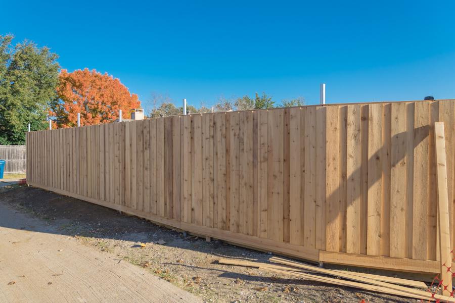 A wooden fence is under construction with fall trees in the background and extra lumber in the foreground. 