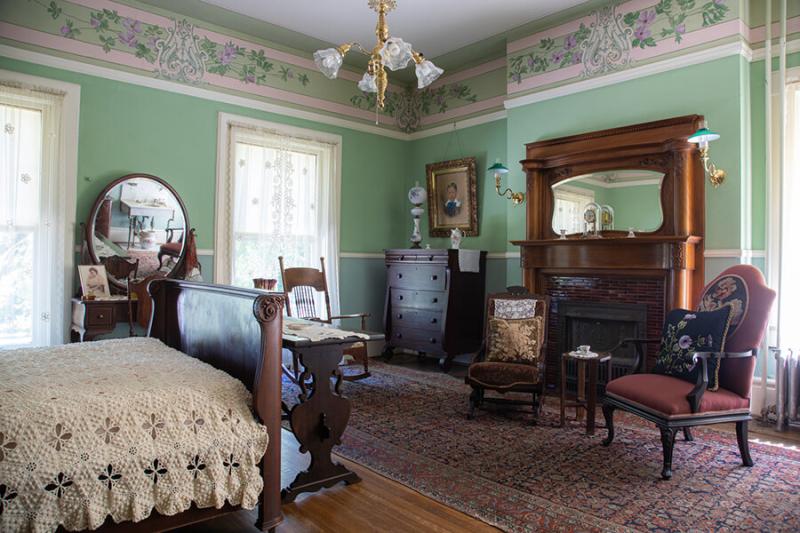 bedroom on the second floor of the Bingham-Waggoner Estate with a bed on the left and a fireplace on the right with two chairs in front of the fireplace. 
