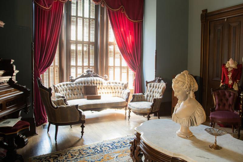 The music and sitting room in the Vaile Mansion in Independence Missouri. Piano is in the left corner with a love seat and two chairs; desk with a bust in the foreground. 