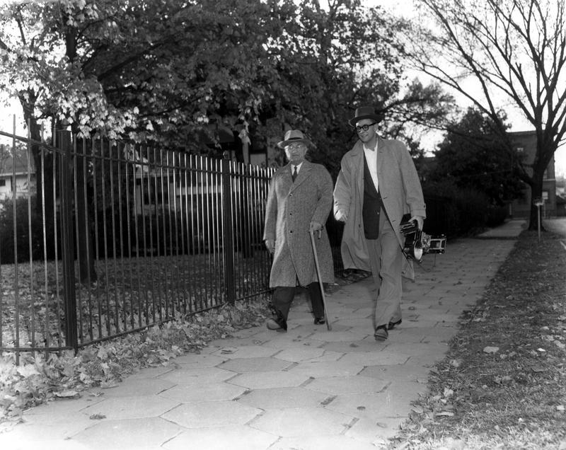 Image of Harry S. Truman walking in his Independence, Missouri neighborhood. Member of the press walks beside him just outside of Truman Home.