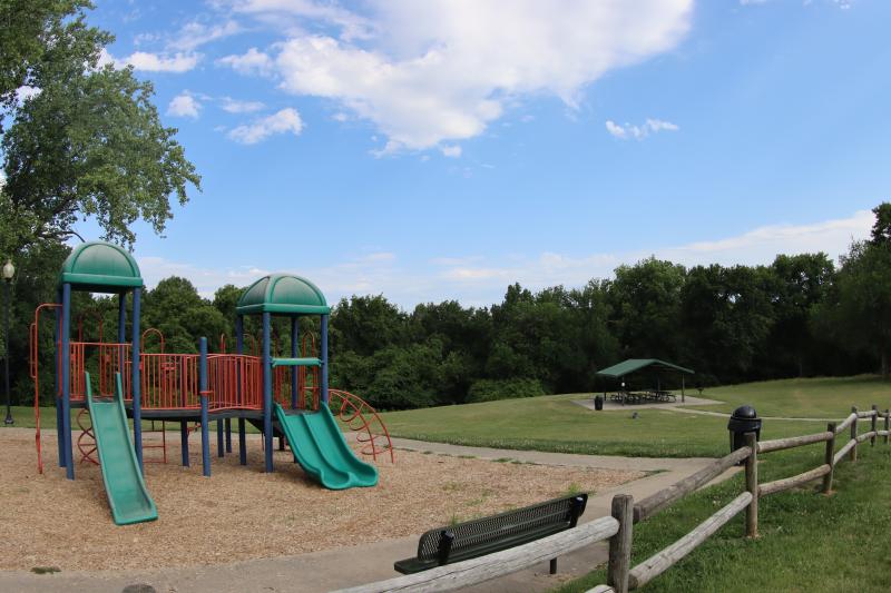 An image of the Dickinson Park playground with a fence in front of it and the park shelter and trees in the background.