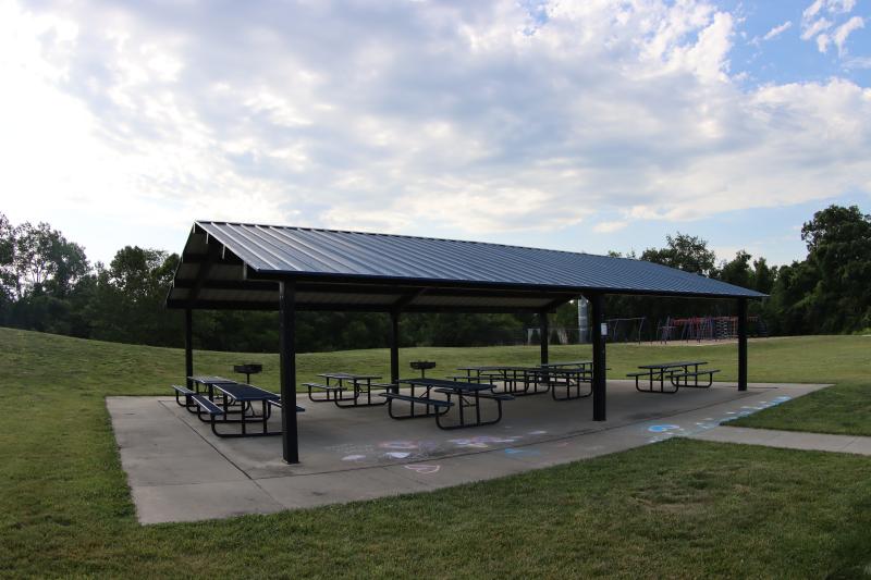 An image of the Independence Athletic Complex pavilion with picnic tables and a few grills on a concrete pad with grass surrounding it on a cloudy day.