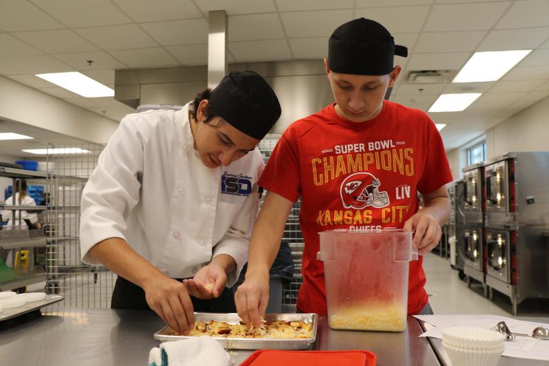 Two students, one in a white chef's coat and another in a red Kansas CIty Chiefs shirt, sprinkle cheese on peta