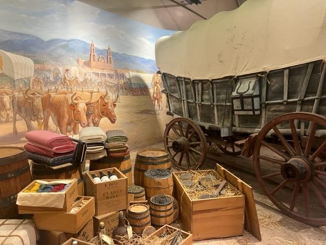 An image of an 1800s wagon with supplies for travel and a backdrop showing pioneers with their wagons preparing to head out West.