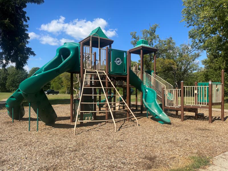 An image of a green, brown and beige colored playground located at Hill Park on a partly cloudy day.