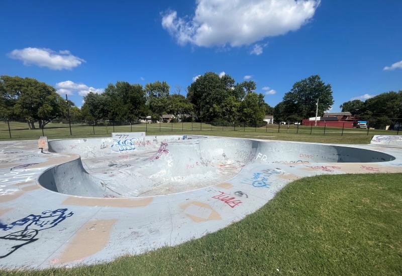 An image of a concrete skate park marked with graffiti located at Hill Park on a partly cloudy day.