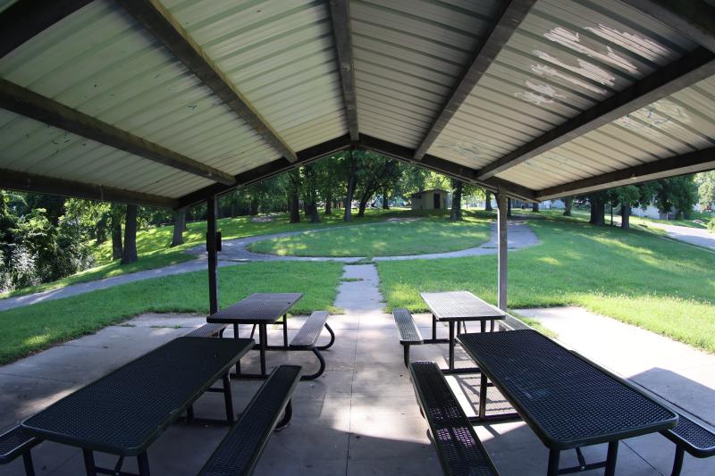 An inside view of the Fairmount Park shelter structure with four picnic tables with a sidewalk leading up to the restroom structure in the background surrounded by trees.