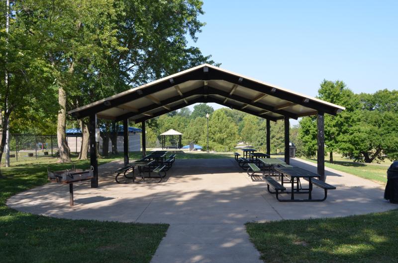 An image of the front view of the McCoy Park pavilion structure with seven picnic tables underneath and a double grill.