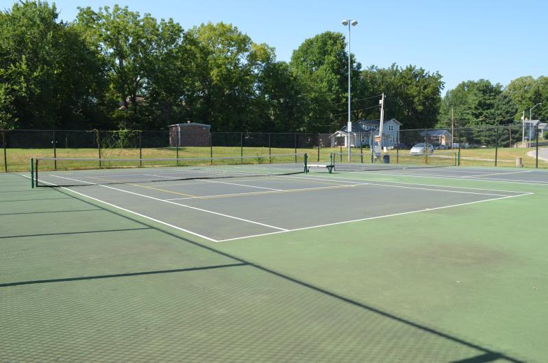 An image of the two tennis courts surrounded by a fence and a court light pole at McCoy Park with trees in the background.