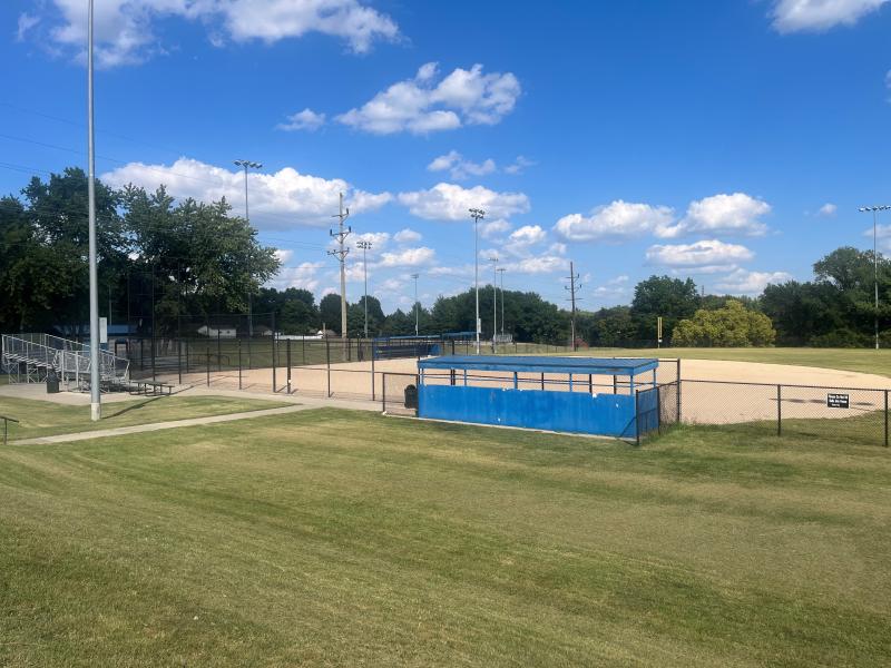 An image of one of the baseball fields with a blue dugout and light poles at Mill Creek Park on a partly cloudy day.