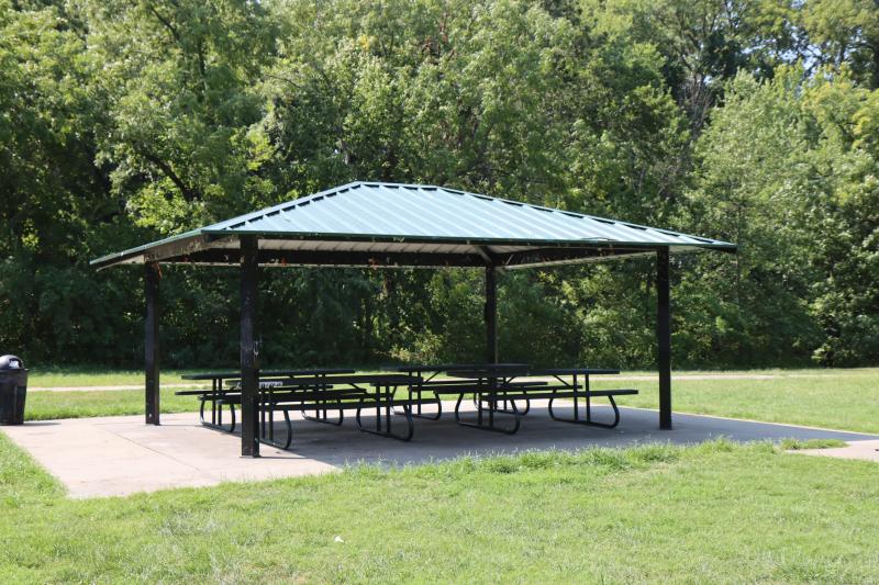 An image of the Rotary Park shelter with picnic tables surrounded by grass and trees in the background.