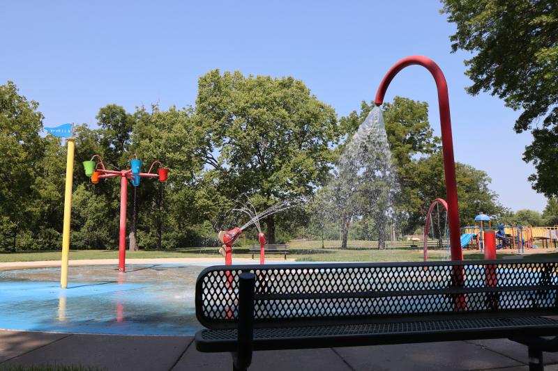 An image of a splash pad area with water spraying out of the water features at Rotary Park with a bench in the shade nearby.