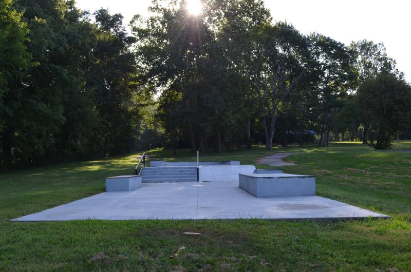 An image of the west side of the concrete skate area located at Santa Fe Park with the sun hiding behind tall trees in the background.