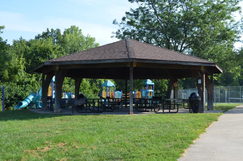 An image of the round pavilion structure with picnic tables underneath at Santa Fe Park. A playground, tennis courts and trees are behind the pavilion.