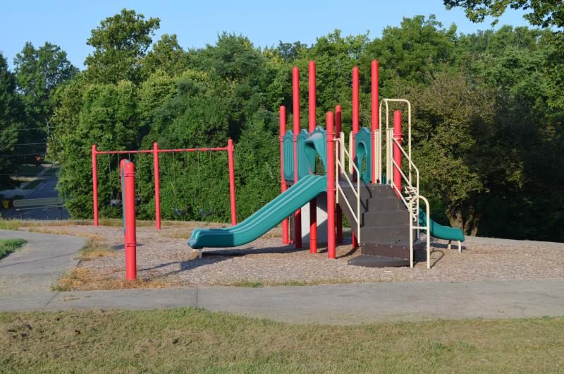 An image of a green and red playground with swings behind it at Van Hook Park. Trees are in the background.