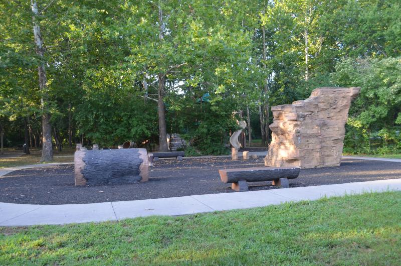 An image of a play area with a few log style benches surrounded by a sidewalk at Waterfall Park. 
