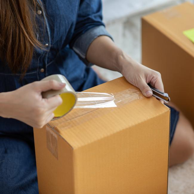 A woman in a denim dress tapes up and labels moving boxes.