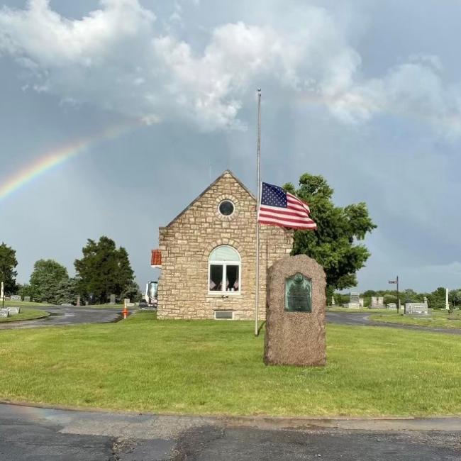 An image of the Woodlawn Cemetery office rock building inside the cemetery grounds with an American flag in front of it and a rainbow in the sky.