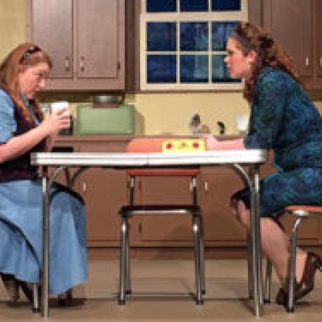 Two women talking across a table while performing on stage at the City Theatre of Independence