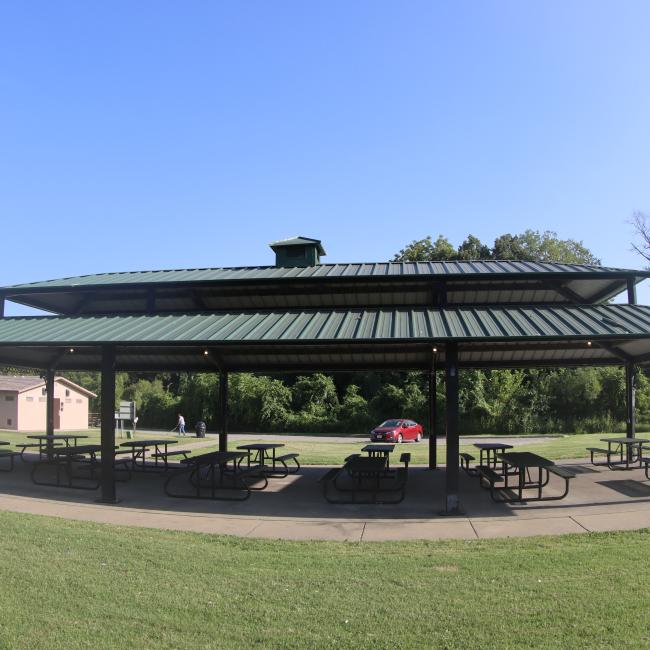 An image of the pavilion structure with 12 picnic tables and 3 grills at Hill Park with the restroom structure in the background.