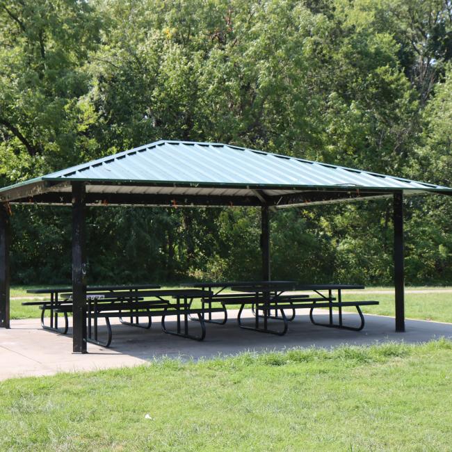 An image of the Rotary Park shelter with picnic tables surrounded by grass and trees in the background.