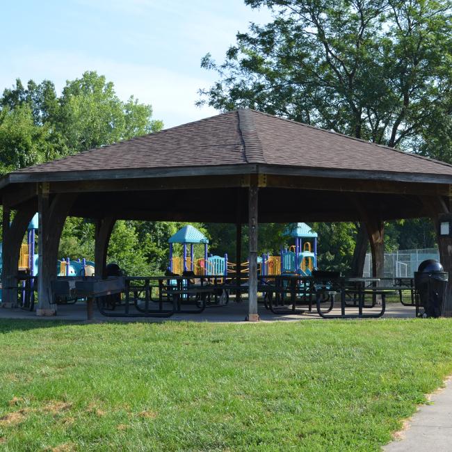 An image of the round pavilion structure with picnic tables underneath at Santa Fe Park. A playground, tennis courts and trees are behind the pavilion.
