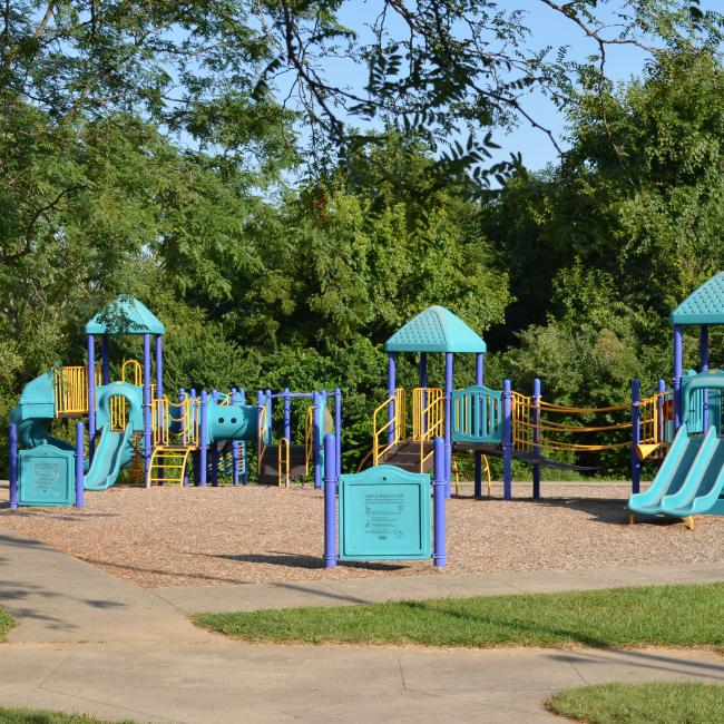 An image of a blue and yellow playground area surrounded by a sidewalk and two benches at Santa Fe Park.