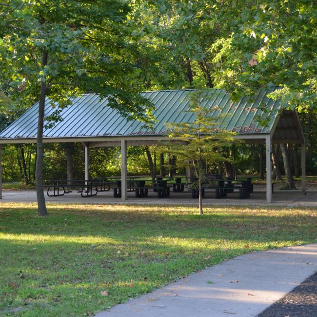A side view of the Waterfall Park pavilion with picnic tables under it surrounded by trees and a sidewalk.